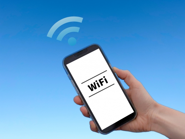 澄み切った青空を背景に、画面に「WiFi」という文字が表示されたスマートフォンを手に持っています。携帯電話の上には、ワイヤレス接続を示す WiFi 信号アイコンが描かれています。.